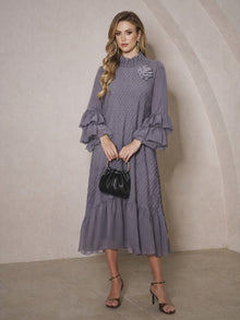 A woman holds a black purse while wearing the Social Queen Floral Appliqué Embroidered Ruffle Dress, featuring long sleeves and delicate details, standing against a beige textured wall.