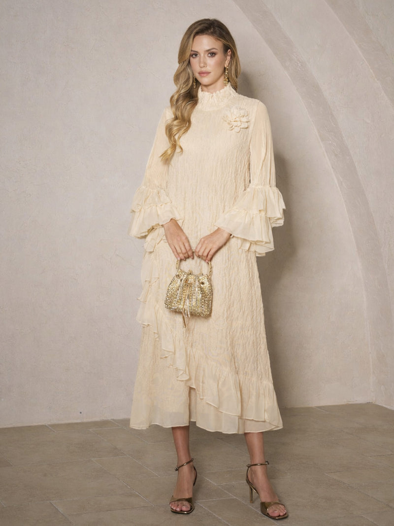 A woman stands indoors wearing the Floral Appliqué Embroidered Ruffle Dress by Social Queen, paired with a small gold handbag, gold earrings, and heeled sandals.