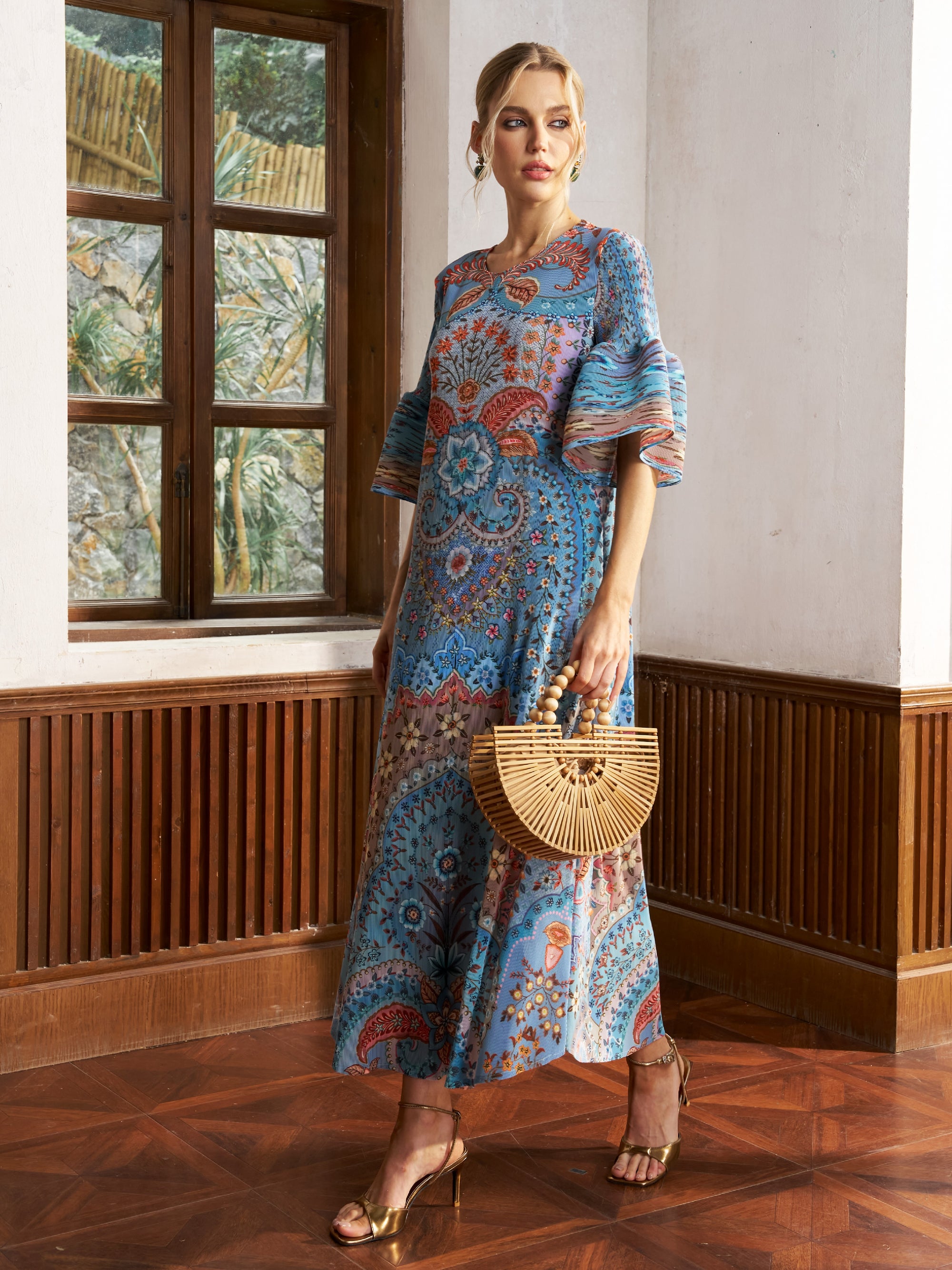 A woman stands indoors in the Social Queen Paisley Print Pleated Midi Dress, paired with gold heels and a bamboo handbag, with a window and wooden paneling in the background.
