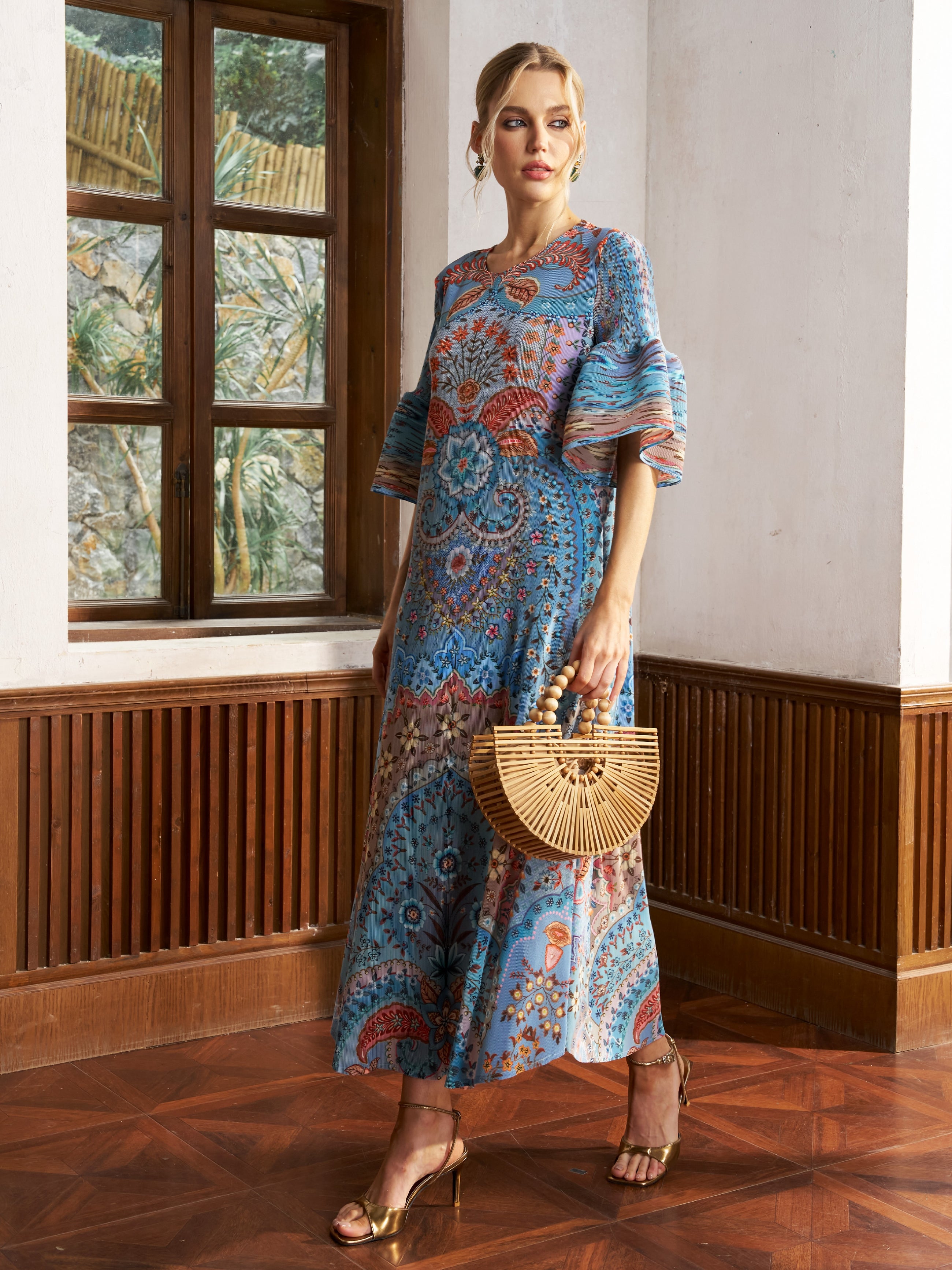 A woman stands indoors in the Social Queen Paisley Print Pleated Midi Dress, paired with gold heels and a bamboo handbag, with a window and wooden paneling in the background.