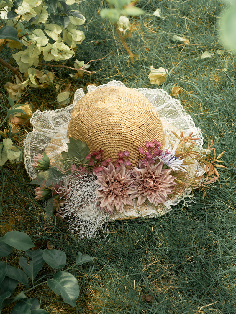 The Social Queen 3D Dahlia Raffia Wide-Brim Hat, adorned with lace, artificial flowers, and leaves, rests on grass amid lush greenery.