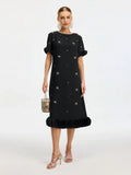 A woman wears the Social Queen Beaded Crystal Embellished Dress, featuring a black, knee-length shift silhouette with star and crystal details, ruffled sleeves and hem. She accessorizes with a small handbag, silver sandals, and statement earrings.