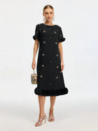 A woman wears the Social Queen Beaded Crystal Embellished Dress, featuring a black, knee-length shift silhouette with star and crystal details, ruffled sleeves and hem. She accessorizes with a small handbag, silver sandals, and statement earrings.