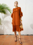 A woman stands on a tiled floor in the Social Queen Beaded Floral Pleated Midi Dress, paired with black heels, statement earrings, and a woven handbag. A green plant is visible in the background.