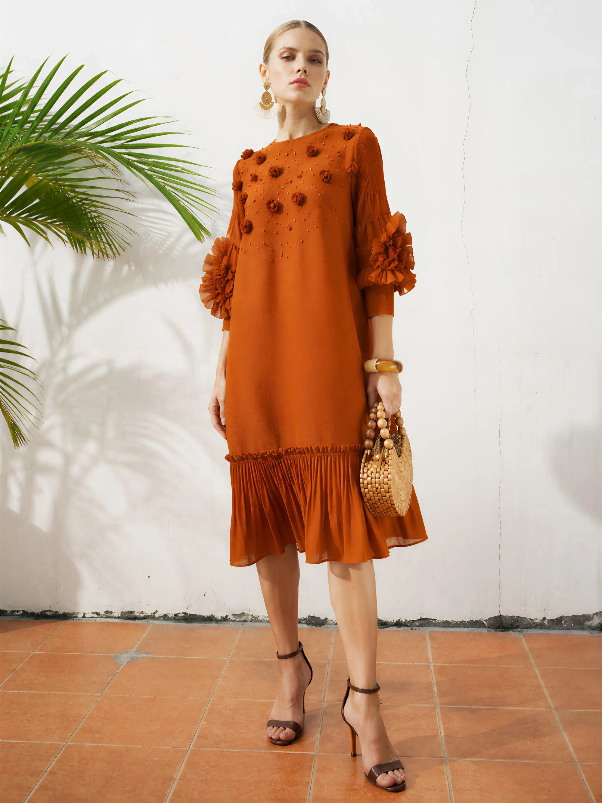 A woman stands on a tiled floor in the Social Queen Beaded Floral Pleated Midi Dress, paired with black heels, statement earrings, and a woven handbag. A green plant is visible in the background.