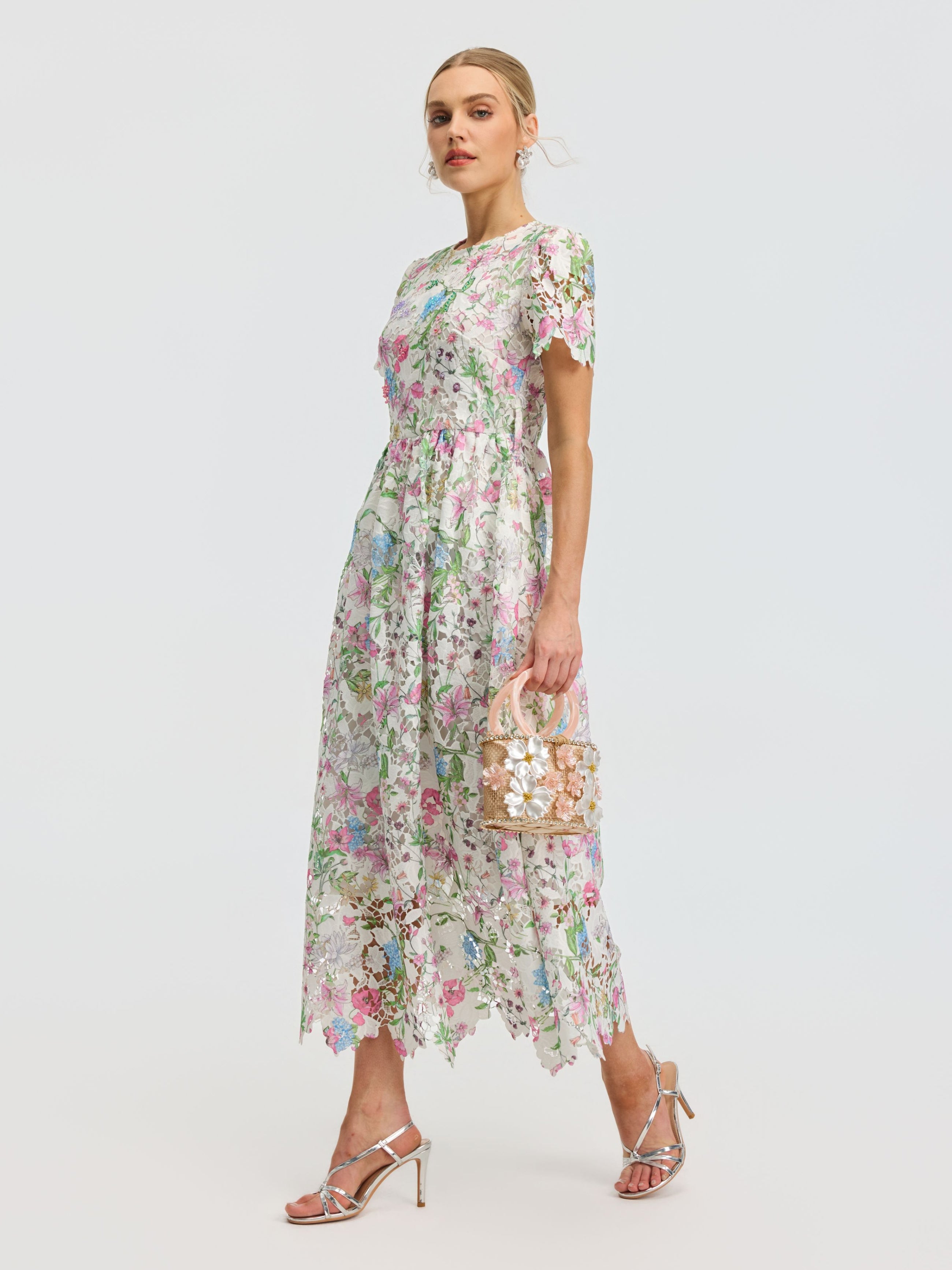 A woman in the Social Queen Botanical Embroidered Cutout Dress and silver heels carries a decorative handbag, walking against a plain light background.