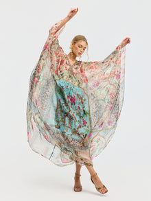 A woman poses with arms raised, wearing the Social Queen Floral Bird Silk Caftan Dress featuring a blue and pink bird and floral print, paired with brown sandals, set against a plain white background.