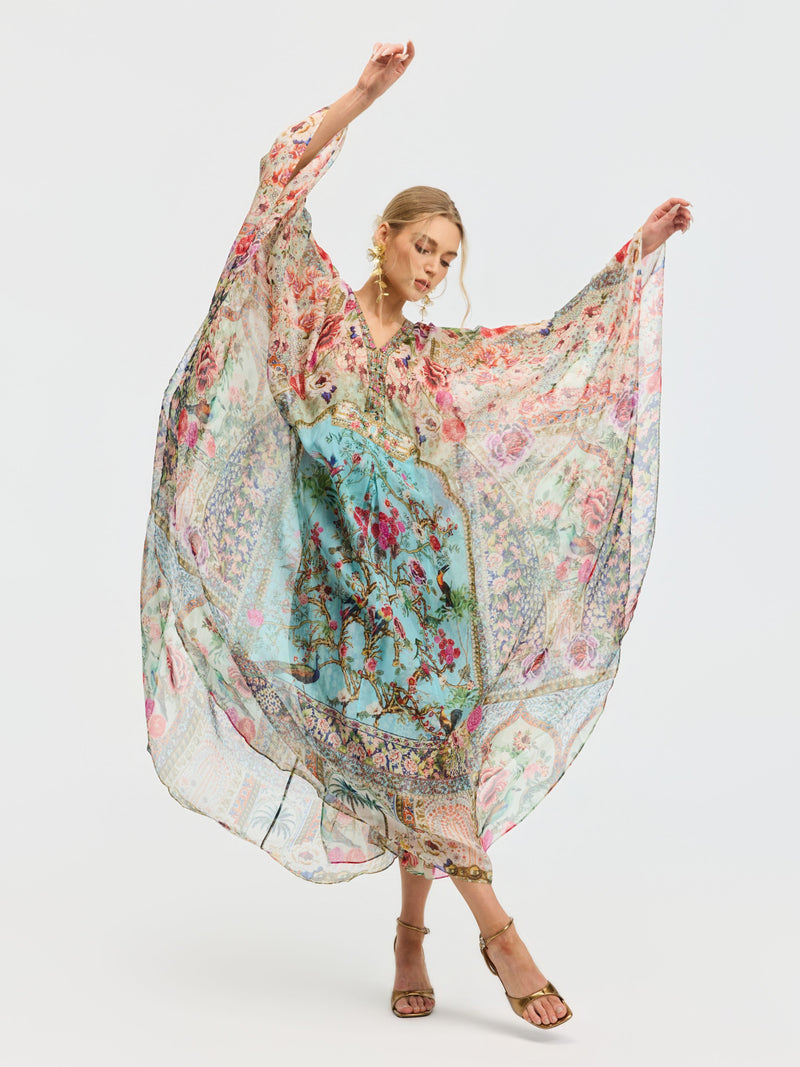 A woman poses with arms raised, wearing the Social Queen Floral Bird Silk Caftan Dress featuring a blue and pink bird and floral print, paired with brown sandals, set against a plain white background.