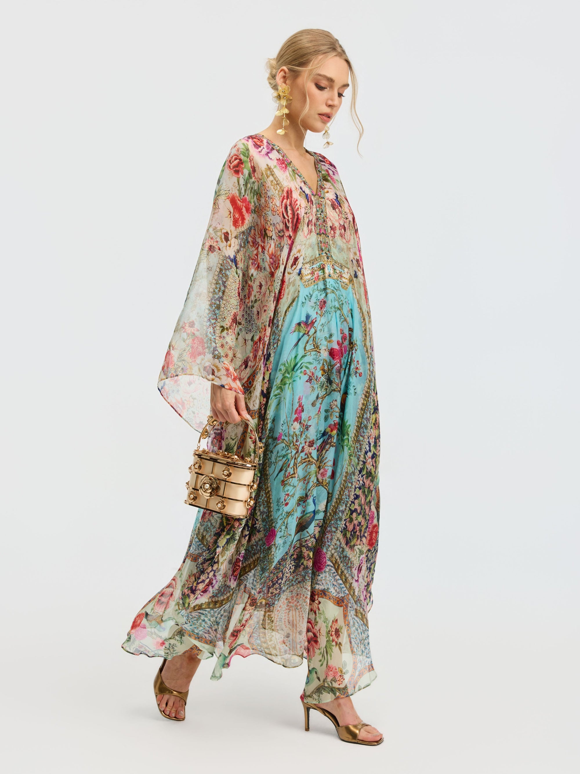 A woman wears the Social Queen Floral Bird Silk Caftan Dress with a multicolored bird and floral print, paired with a round handbag and gold sandals against a plain background.