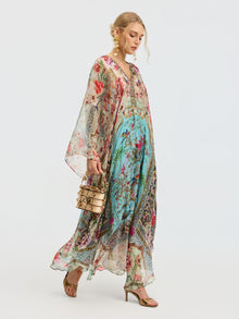 A woman wears the Social Queen Floral Bird Silk Caftan Dress with a multicolored bird and floral print, paired with a round handbag and gold sandals against a plain background.