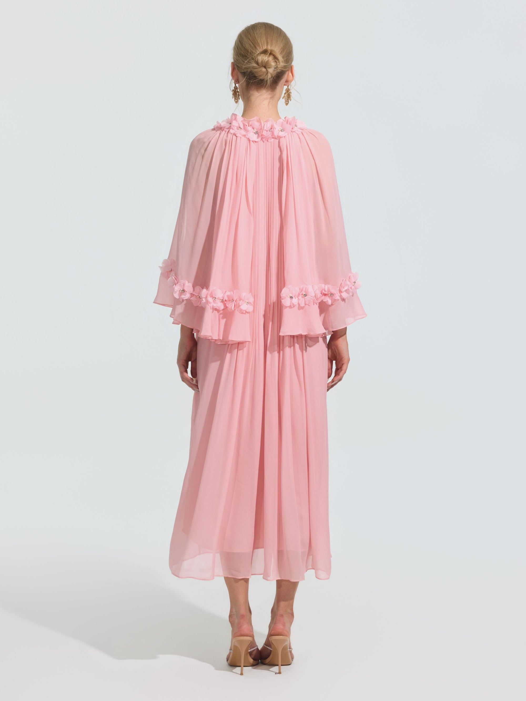 A person stands facing away in the Social Queen 3D Floral Embellished Silk Dress with a flowing pink silhouette, sheer cape sleeves, and floral appliqué, styled with beige heeled sandals and hair in a neat bun.