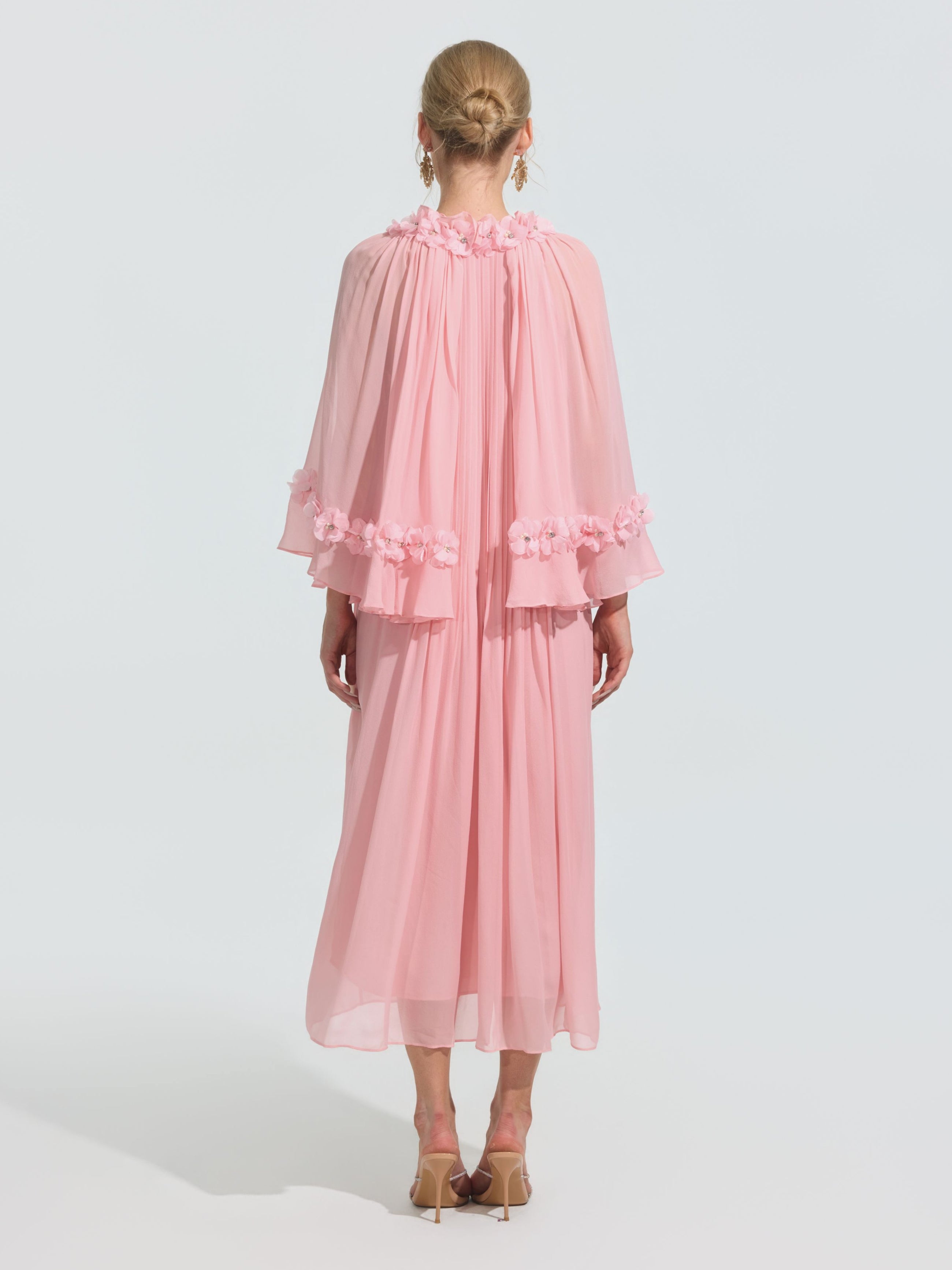 A person stands facing away in the Social Queen 3D Floral Embellished Silk Dress with a flowing pink silhouette, sheer cape sleeves, and floral appliqué, styled with beige heeled sandals and hair in a neat bun.