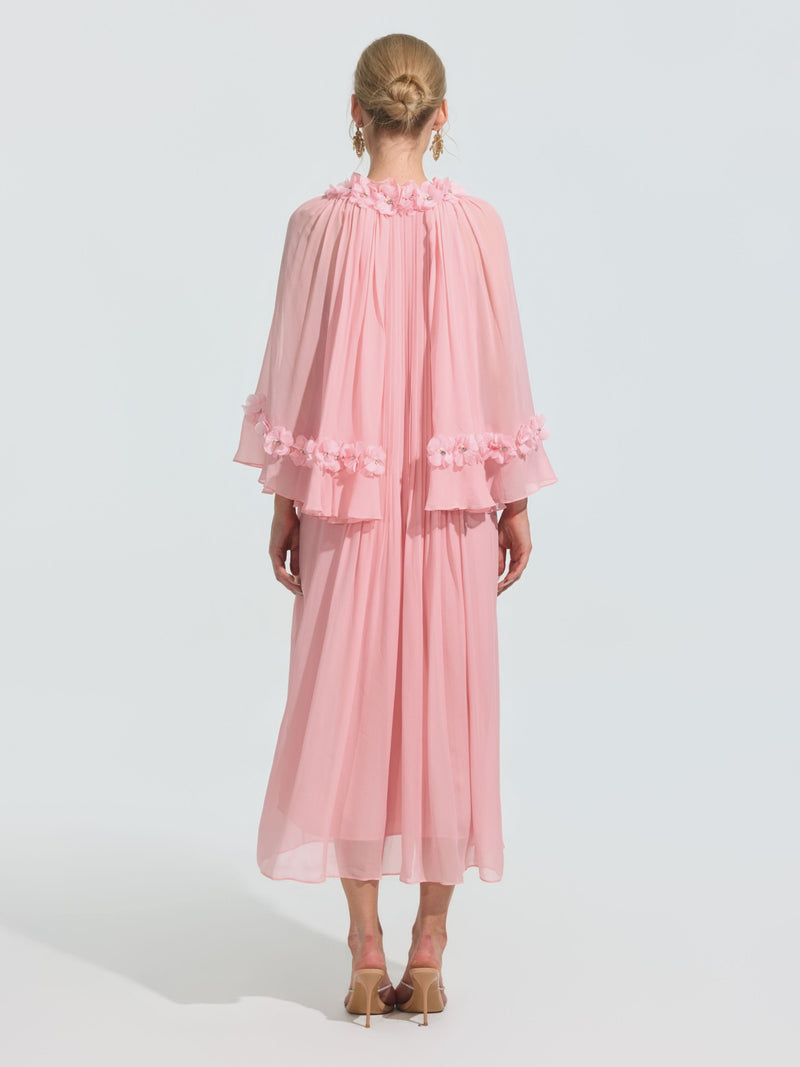 A person stands facing away in the Social Queen 3D Floral Embellished Silk Dress with a flowing pink silhouette, sheer cape sleeves, and floral appliqué, styled with beige heeled sandals and hair in a neat bun.