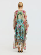 A woman faces away, wearing the Social Queen Floral Bird Silk Caftan Dress with a vibrant print and green bow, paired with gold sandals. Her hair is styled in a neat bun—perfect for elegant resort wear.