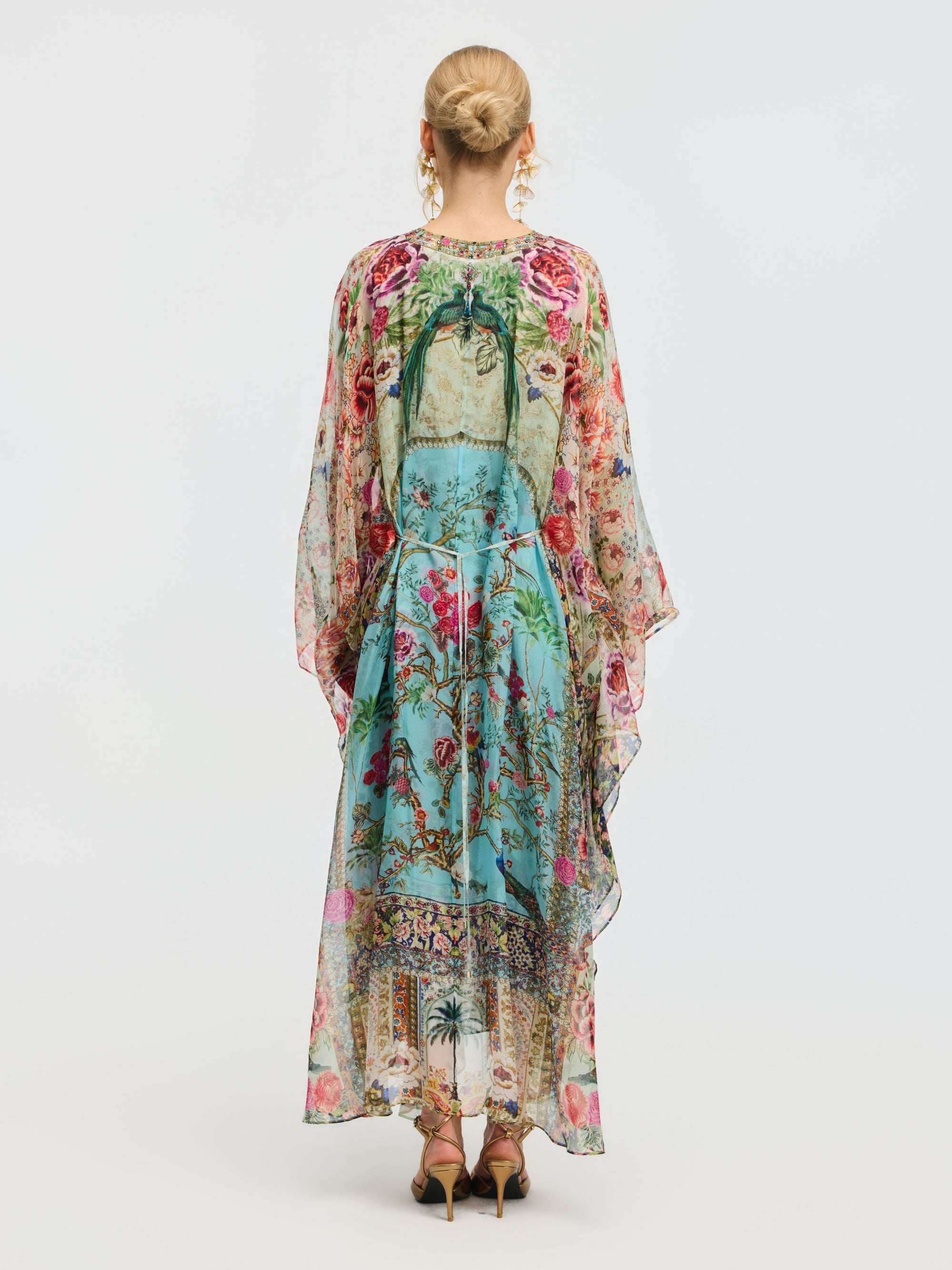 A woman faces away, wearing the Social Queen Floral Bird Silk Caftan Dress with a vibrant print and green bow, paired with gold sandals. Her hair is styled in a neat bun—perfect for elegant resort wear.