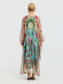 A woman faces away, wearing the Social Queen Floral Bird Silk Caftan Dress with a vibrant print and green bow, paired with gold sandals. Her hair is styled in a neat bun—perfect for elegant resort wear.