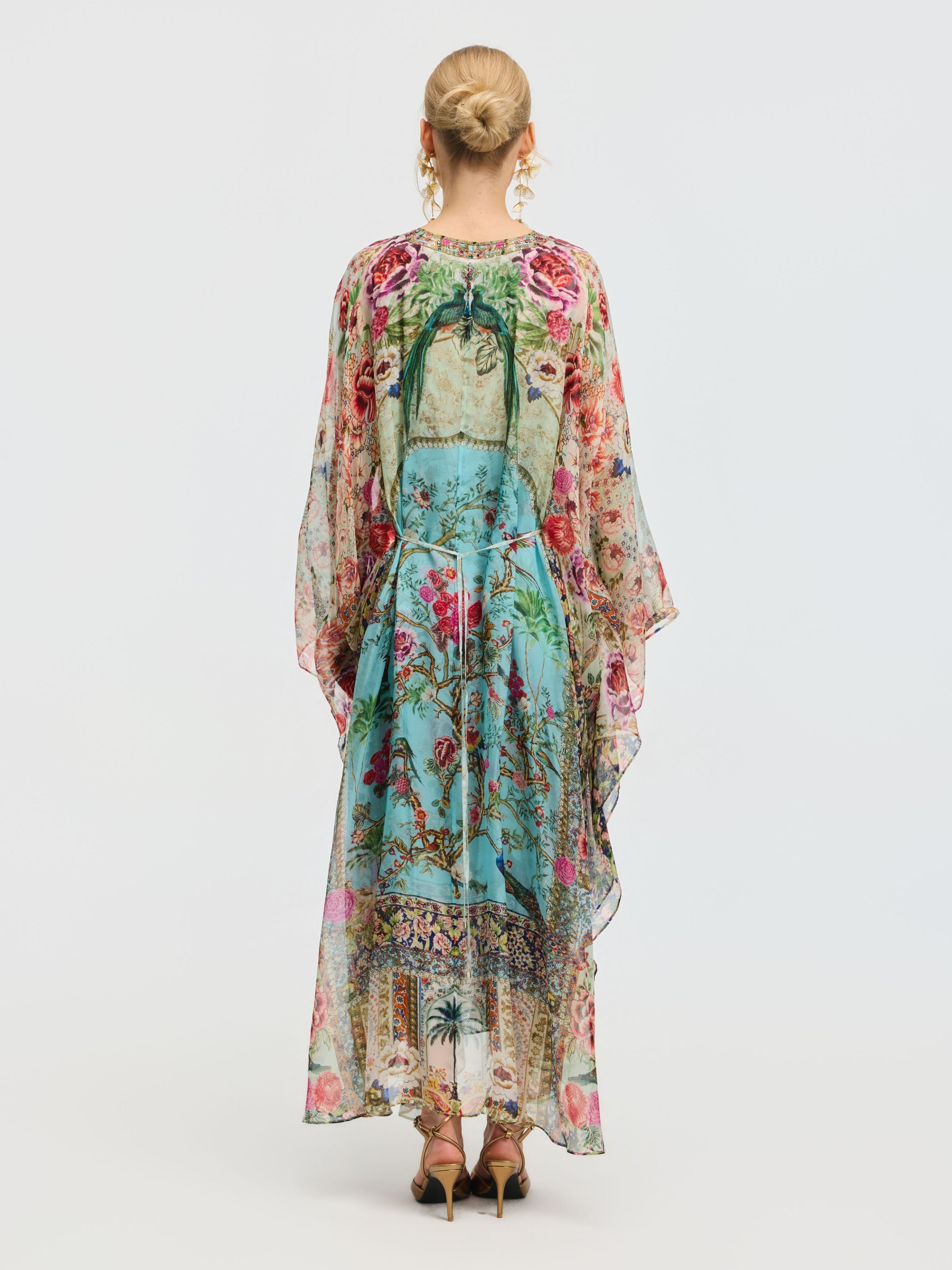 A woman faces away, wearing the Social Queen Floral Bird Silk Caftan Dress with a vibrant print and green bow, paired with gold sandals. Her hair is styled in a neat bun—perfect for elegant resort wear.