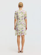A woman faces away, wearing the Social Queen Dimensional Embroidered Beaded Lace Dress featuring a knee-length floral design in white, green, and purple. Her hair is in a bun and she finishes the look with white heeled sandals.