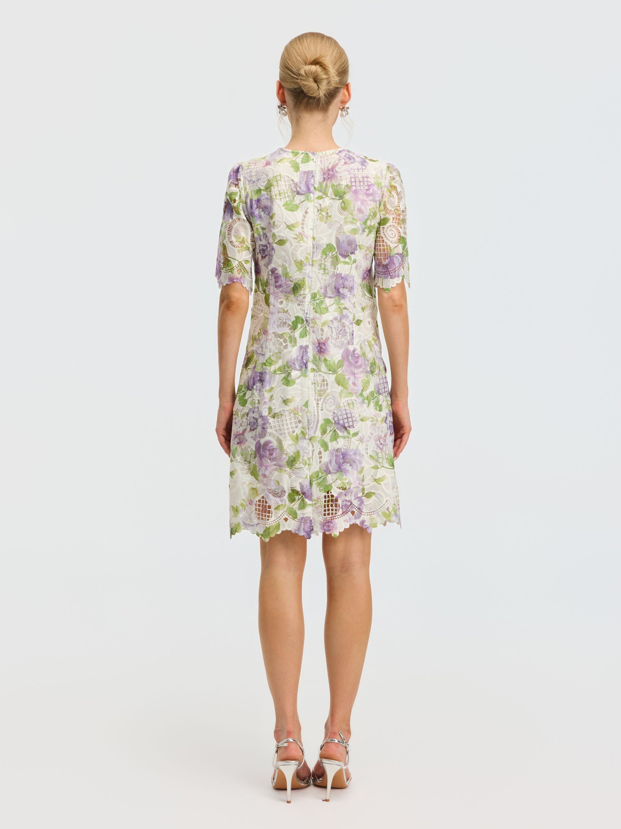 A woman faces away, wearing the Social Queen Dimensional Embroidered Beaded Lace Dress featuring a knee-length floral design in white, green, and purple. Her hair is in a bun and she finishes the look with white heeled sandals.