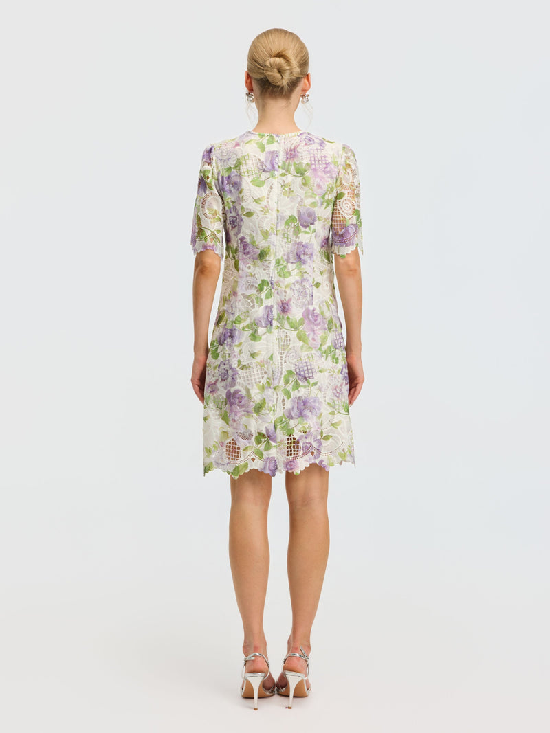 A woman stands facing away, wearing the Social Queen Dimensional Embroidered Beaded Lace Dress in white, green, and purple tones with knee-length, short sleeves, multi-layer lace and white high-heeled sandals. Her hair is styled in a bun.