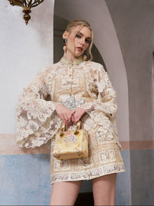 A woman wearing the Social Queen Baroque Print Floral Appliqué Dress—a cream mini dress adorned with intricate floral lace appliqués—stands indoors, holding a small yellow handbag with a floral pattern in front of a textured wall and archway.