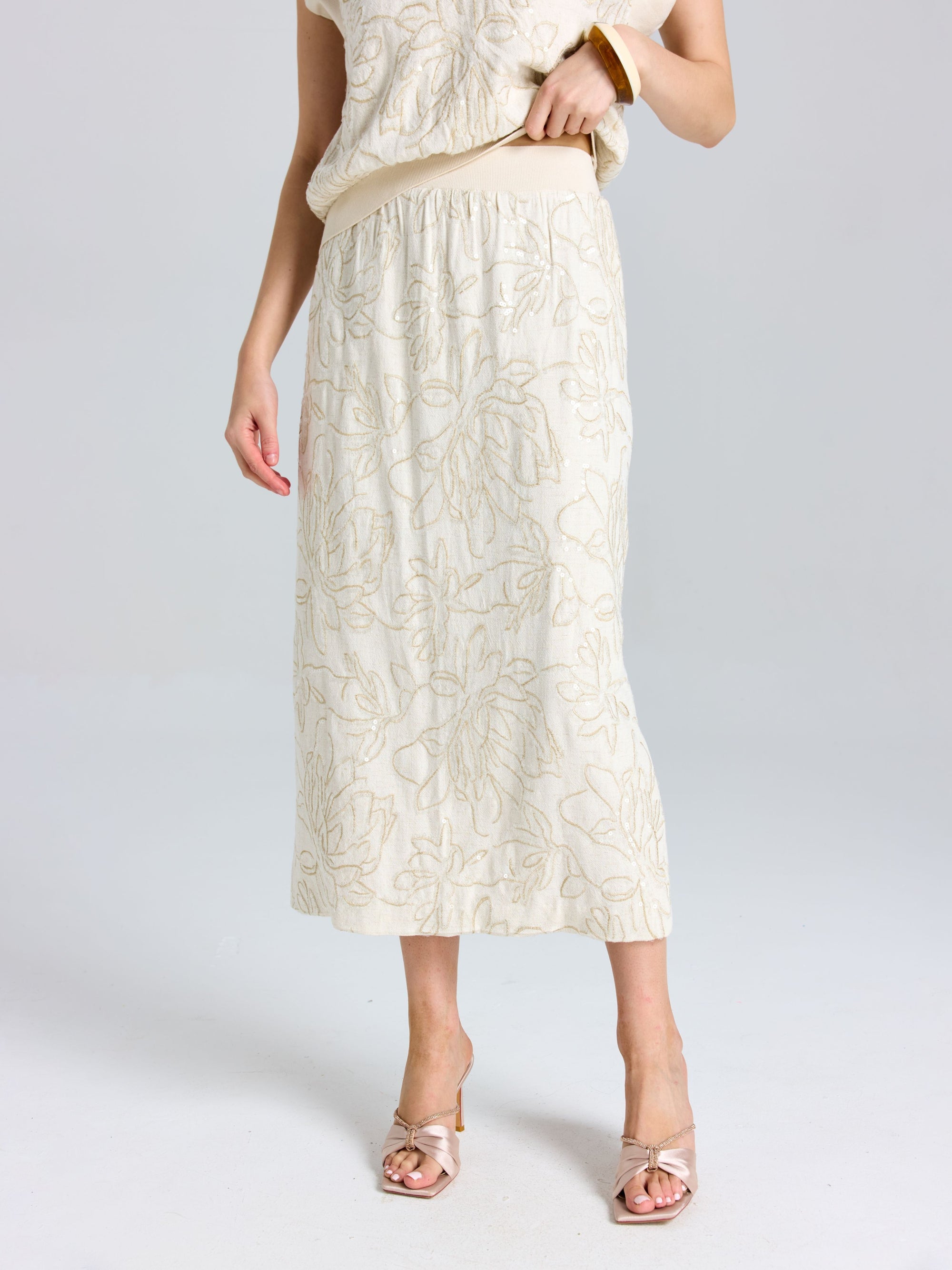 A person models the Social Queen Floral Embroidered Linen Midi Skirt, featuring a cream hue and subtle floral details, styled with open-toe heels against a plain background.