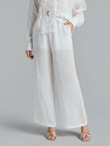 Wearing Social Queen’s Textured Straight-Leg Pants in sheer white, styled with a white lace blouse and metallic open-toe heels, the person stands against a plain background with one hand in their pocket.