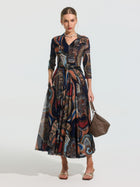 A woman wears the Social Queen Paisley Print Knit Dress (Belt Included), featuring long sleeves and a colorful paisley pattern, paired with sandals and a brown woven handbag.