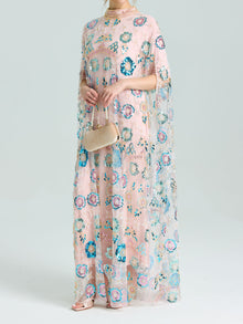 A woman wears the Social Queen Beaded Floral Embellished Cape Gown and holds a small beige handbag, pairing the look with open-toed shoes.