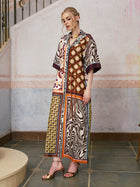A woman stands indoors against a textured wall and tiled floor, wearing the Social Queen Patchwork Print Cotton Shirt Dress (belt included), which features a vibrant patchwork design with short sleeves, paired with gold open-toe heels.