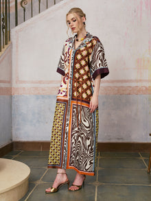A woman stands indoors against a textured wall and tiled floor, wearing the Social Queen Patchwork Print Cotton Shirt Dress (belt included), which features a vibrant patchwork design with short sleeves, paired with gold open-toe heels.
