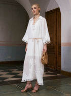A woman stands indoors against wooden doors and a tiled floor, wearing the Social Queen Floral Cutwork Embroidered Linen Dress—a long white linen maxi dress featuring intricate floral cutwork embroidery. She pairs it with gold heels and carries a woven handbag.