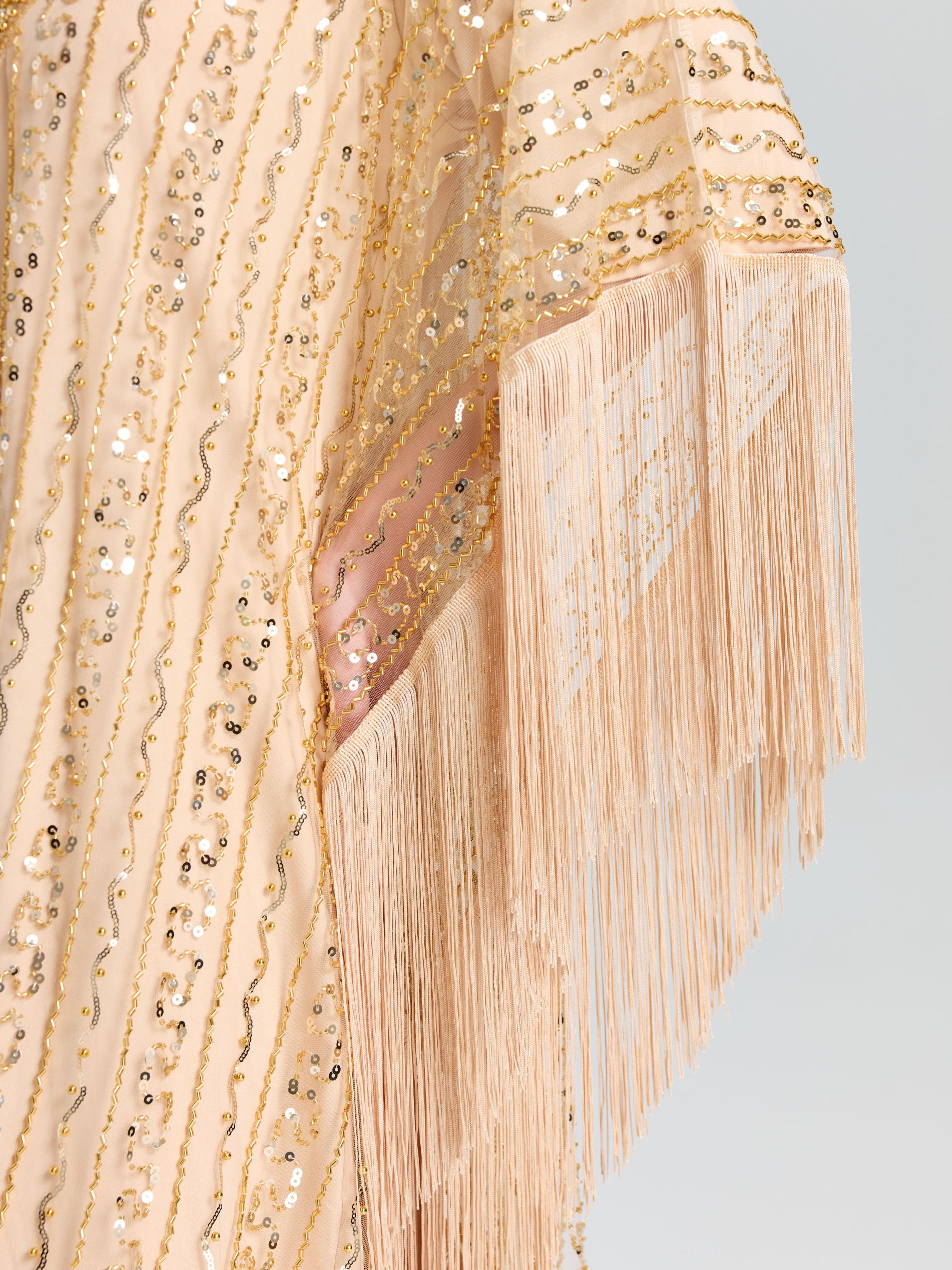 Close-up of the Beaded Fringe Cape Dress by Social Queen, featuring peach fabric with gold sequin waves and long fringe trim—perfect for an elegant look.