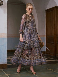 A woman stands indoors on a tiled floor near wooden doors, holding a woven handbag and wearing the Social Queen Leopard Paisley Print Silk Dress—a long, mulberry silk dress combining paisley motifs with leopard print.