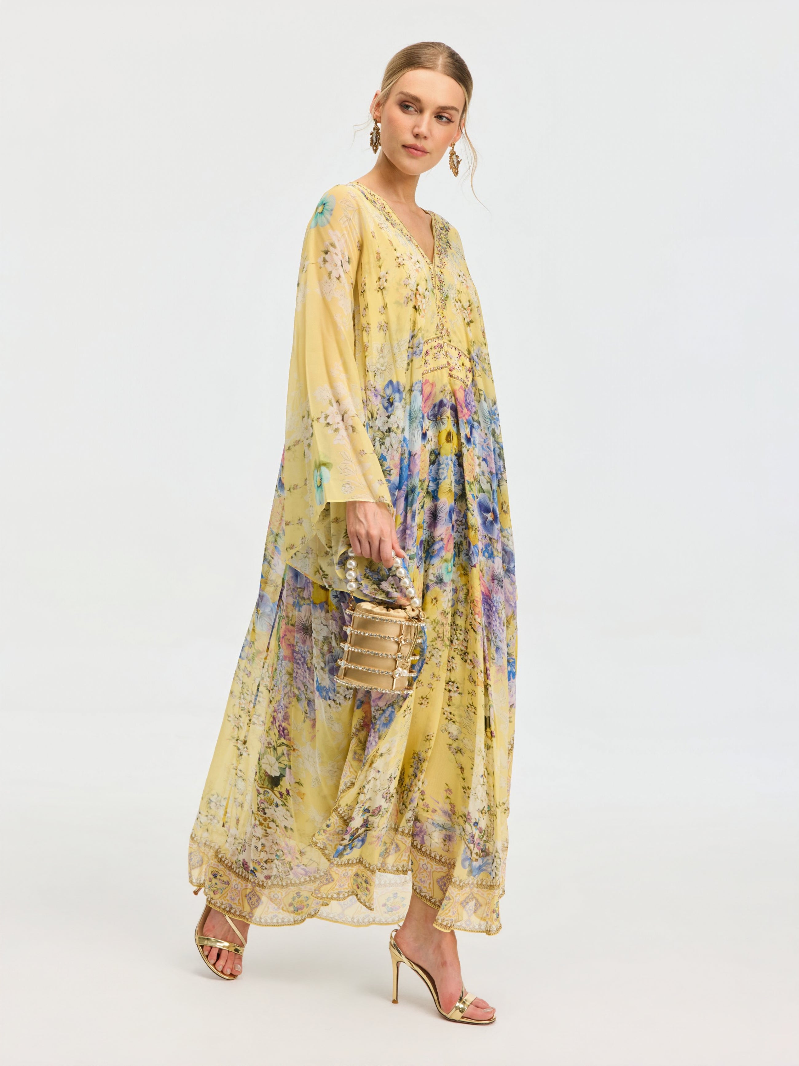 A woman in the Social Queen Floral Print Silk Caftan Dress with gold heels and a beige handbag stands against a plain backdrop—an ideal resort look.
