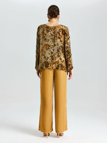 A woman stands with her back to the camera in a plain white studio, wearing Social Queen's Flocked Floral Silk Sheer Top (Camisole Not Included) styled with mustard wide-leg pants.