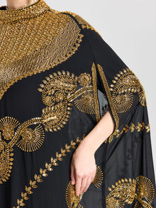A person wears the Social Queen Beaded Embroidered Cape Dress, a black chiffon outfit with gold bead embroidery and Persian-inspired motifs, featuring a high neckline and arm slit where their right hand rests nearby.