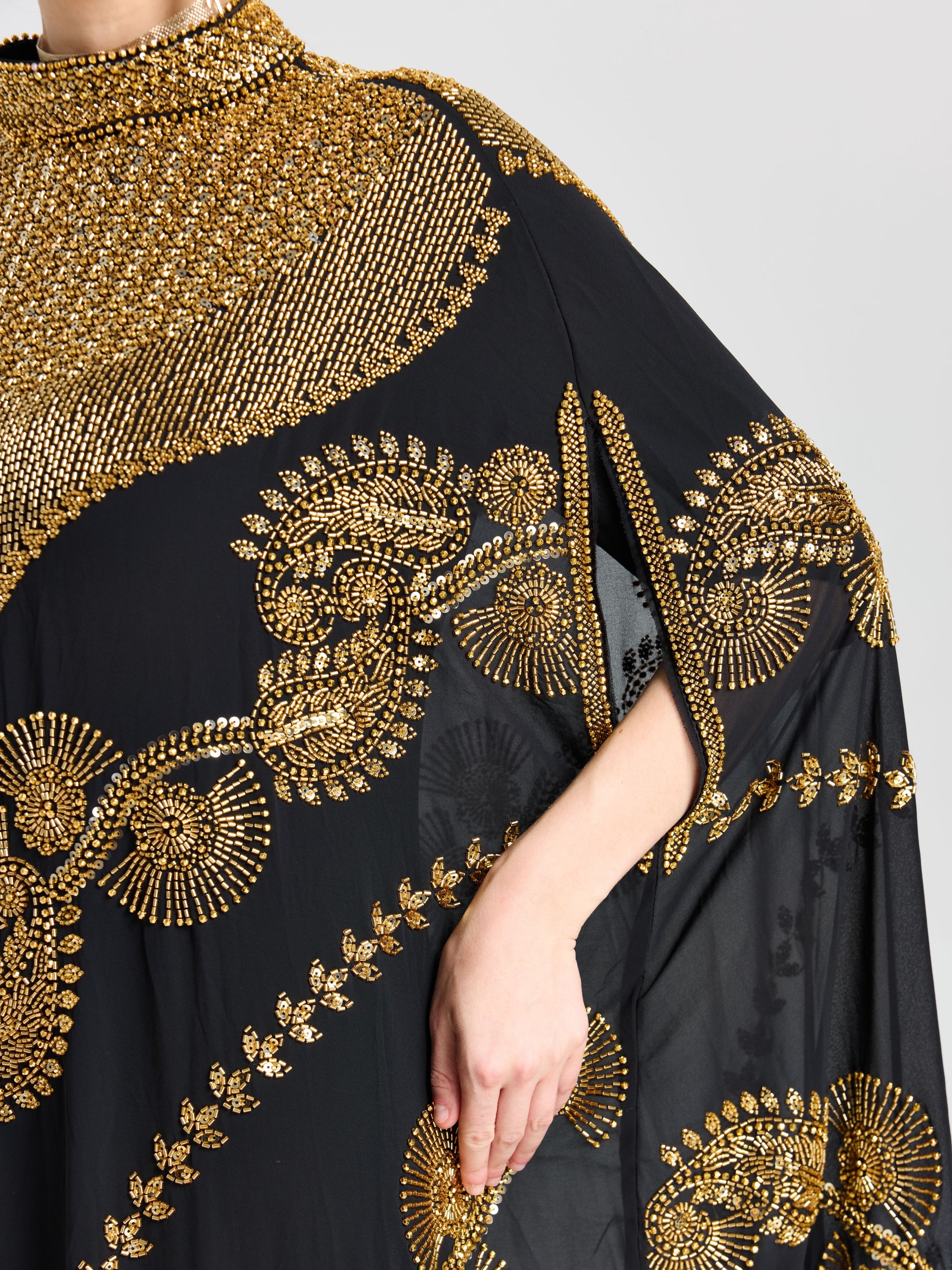 A person wears the Social Queen Beaded Embroidered Cape Dress, a black chiffon outfit with gold bead embroidery and Persian-inspired motifs, featuring a high neckline and arm slit where their right hand rests nearby.