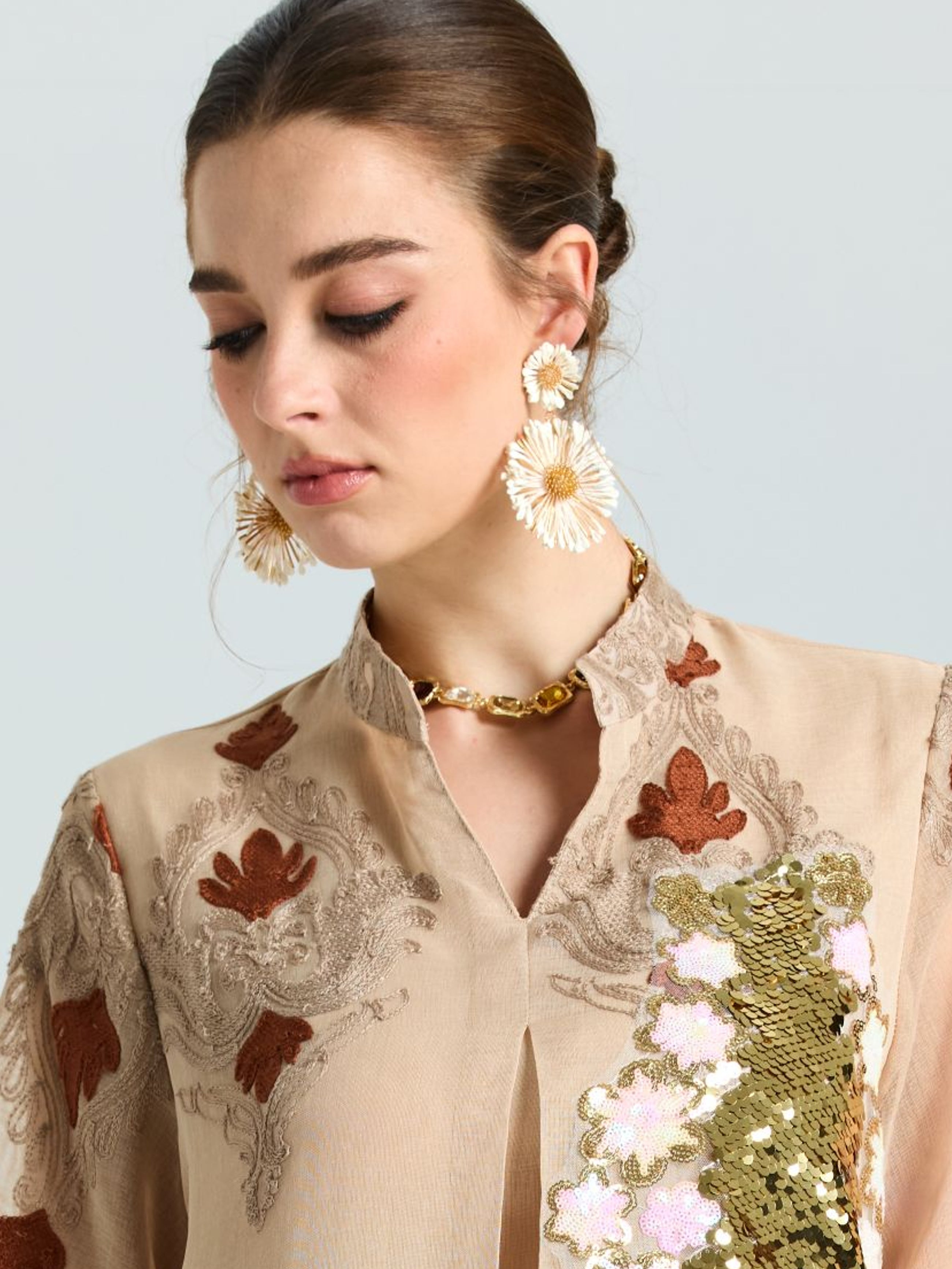 A woman wearing a beige embroidered top with gold and white sequin details, a gold chain necklace, and Social Queen’s Daisy Cluster Earrings, looks down against a plain background.