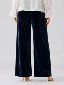 A person is shown from the back wearing Social Queen’s Silk Velvet Wide-Leg Pants in navy blue, paired with a white long-sleeve blouse featuring ruffled cuffs and beige heeled sandals.