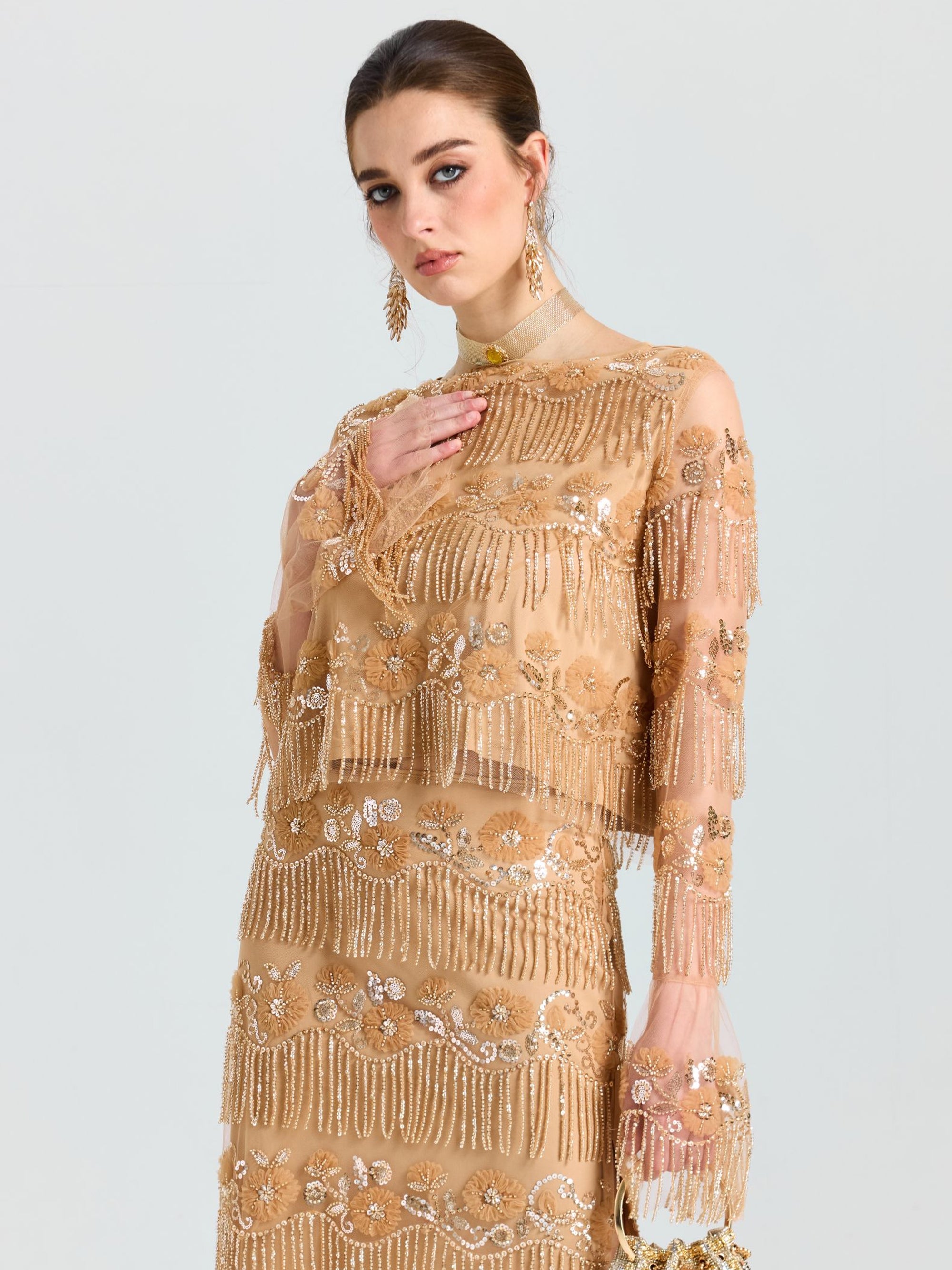 A woman models the Social Queen Beaded Fringe Floral Top, featuring gold-beige sheer fabric with sequins, beaded fringe, and floral embroidery, resting her hand on her chest against a plain light background.