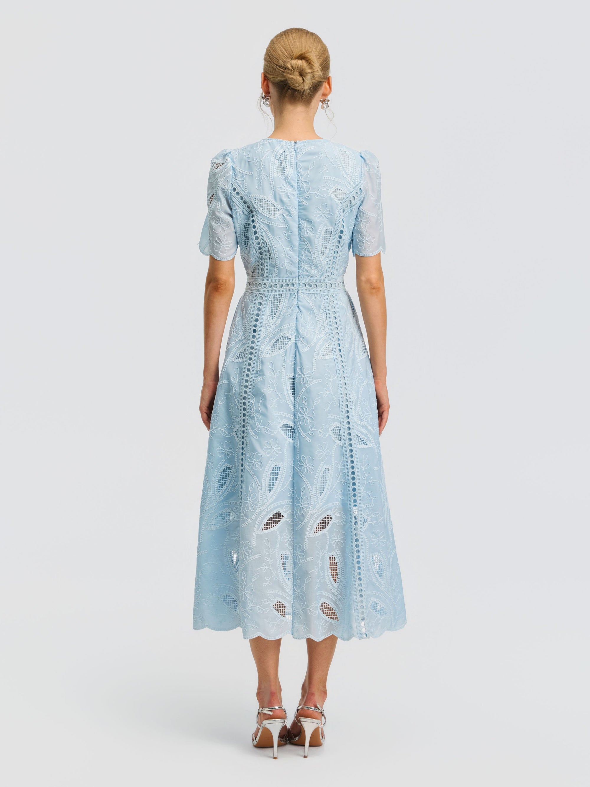 A woman stands with her back to the camera, wearing the Social Queen Floral Cutwork Embroidered Dress—a light blue, knee-length dress adorned with delicate floral embroidery and short sleeves—paired with white heeled sandals, set against a plain white background.