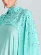 A woman wears the Social Queen Beaded Cape Scarf Gown in aqua blue, featuring sequin details and long dangling earrings. Only her lower face and upper body are visible.