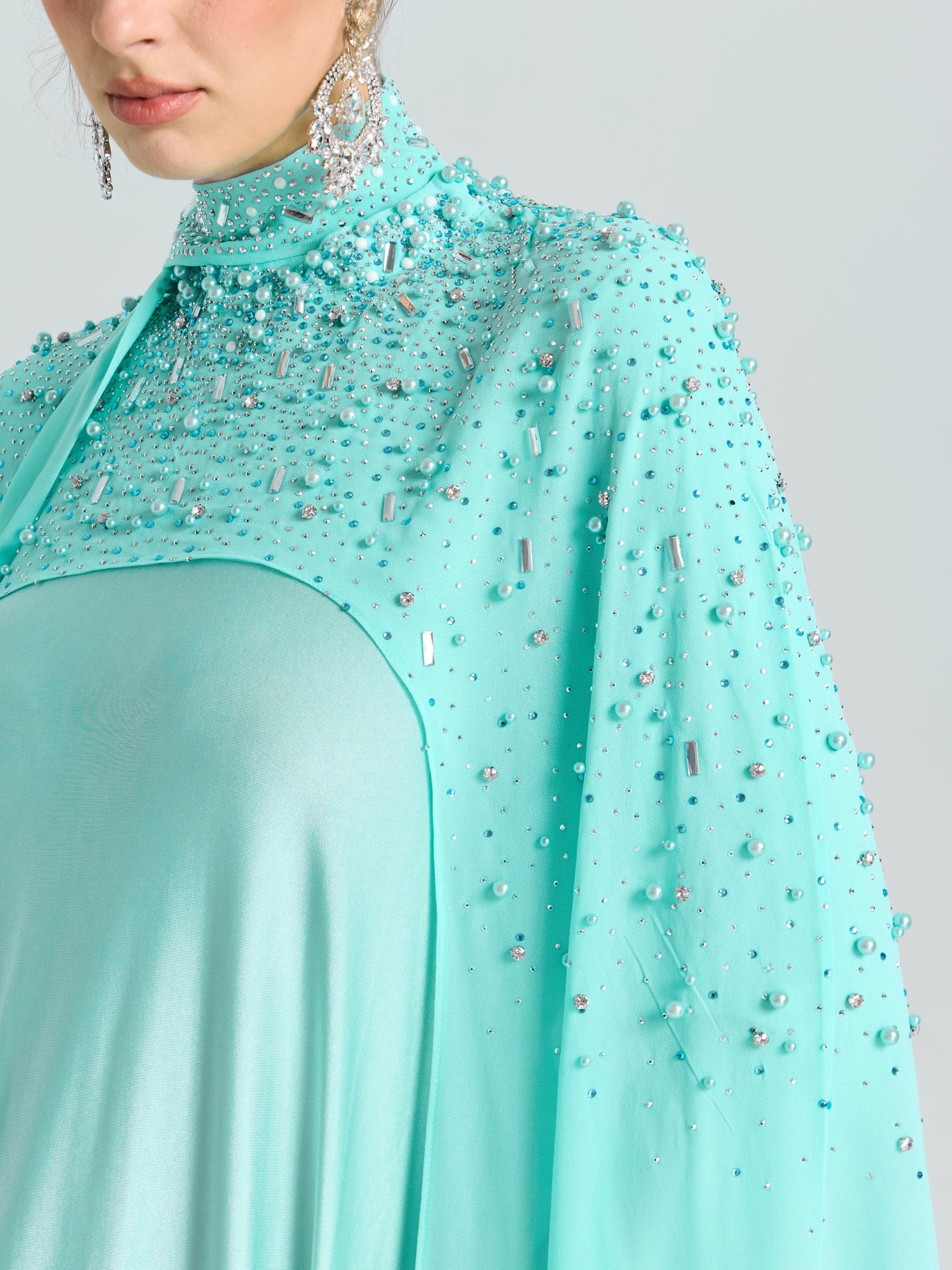A woman wears the Social Queen Beaded Cape Scarf Gown in aqua blue, featuring sequin details and long dangling earrings. Only her lower face and upper body are visible.