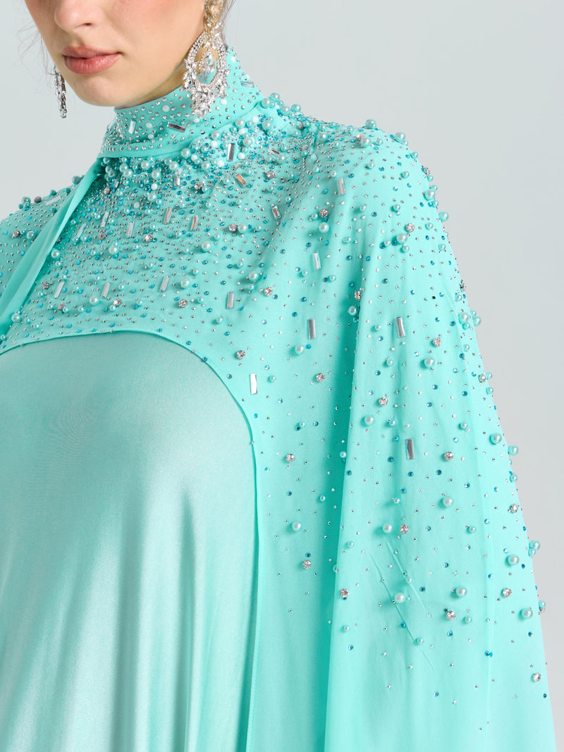 A woman wears the Social Queen Beaded Cape Scarf Gown in aqua blue, featuring sequin details and long dangling earrings. Only her lower face and upper body are visible.