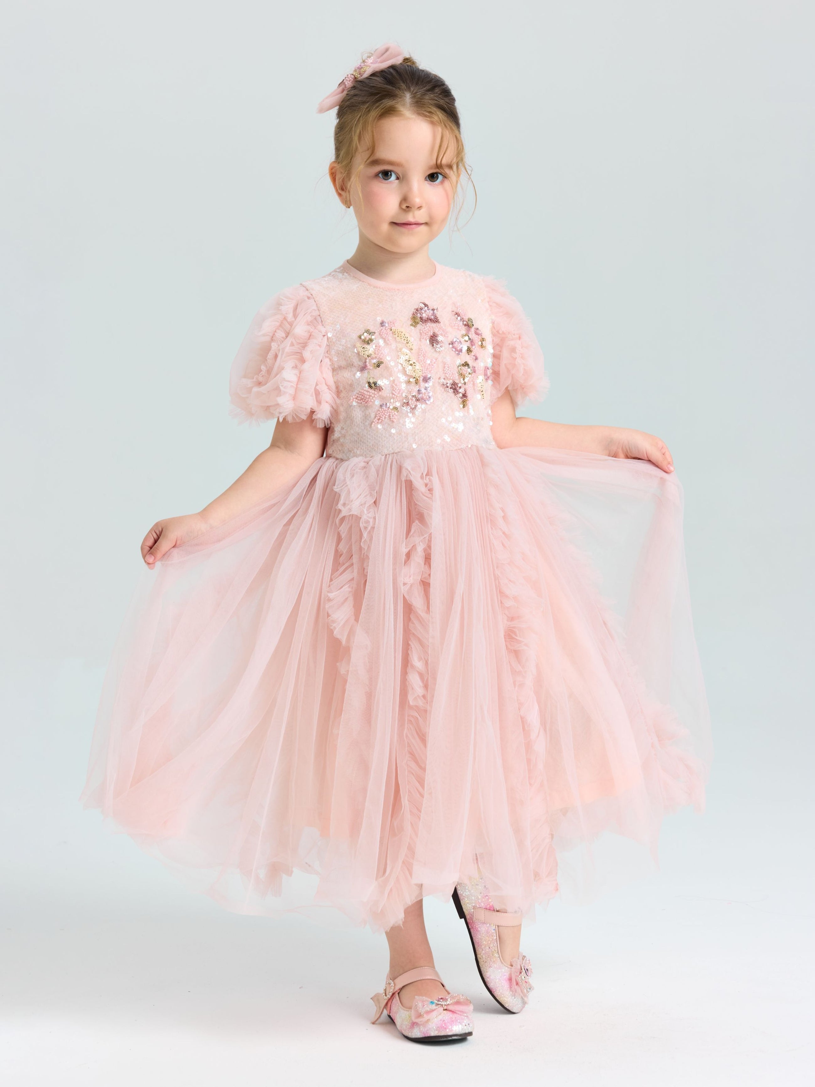 A young girl wears the Social Queen Girls Sequin Embellished Tulle Dress, featuring a light pink princess design with floral details, matching shoes, and a bow in her hair, posing against a plain light background.