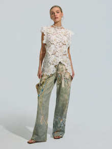 A woman stands wearing the Social Queen Pearl Rose Guipure Lace Top, paired with wide-leg, light-wash jeans adorned with a decorative pattern and subtle pearl details, and carries a small handbag.
