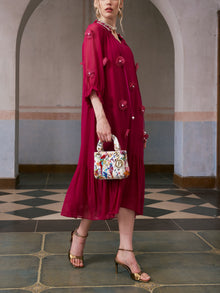 A woman wearing the Social Queen 3D Floral Pearl Mulberry Silk Dress—a loose, knee-length magenta piece adorned with 3D floral appliqué—pairs it with gold high heels and carries a small, white patterned handbag while standing indoors on a tiled floor.