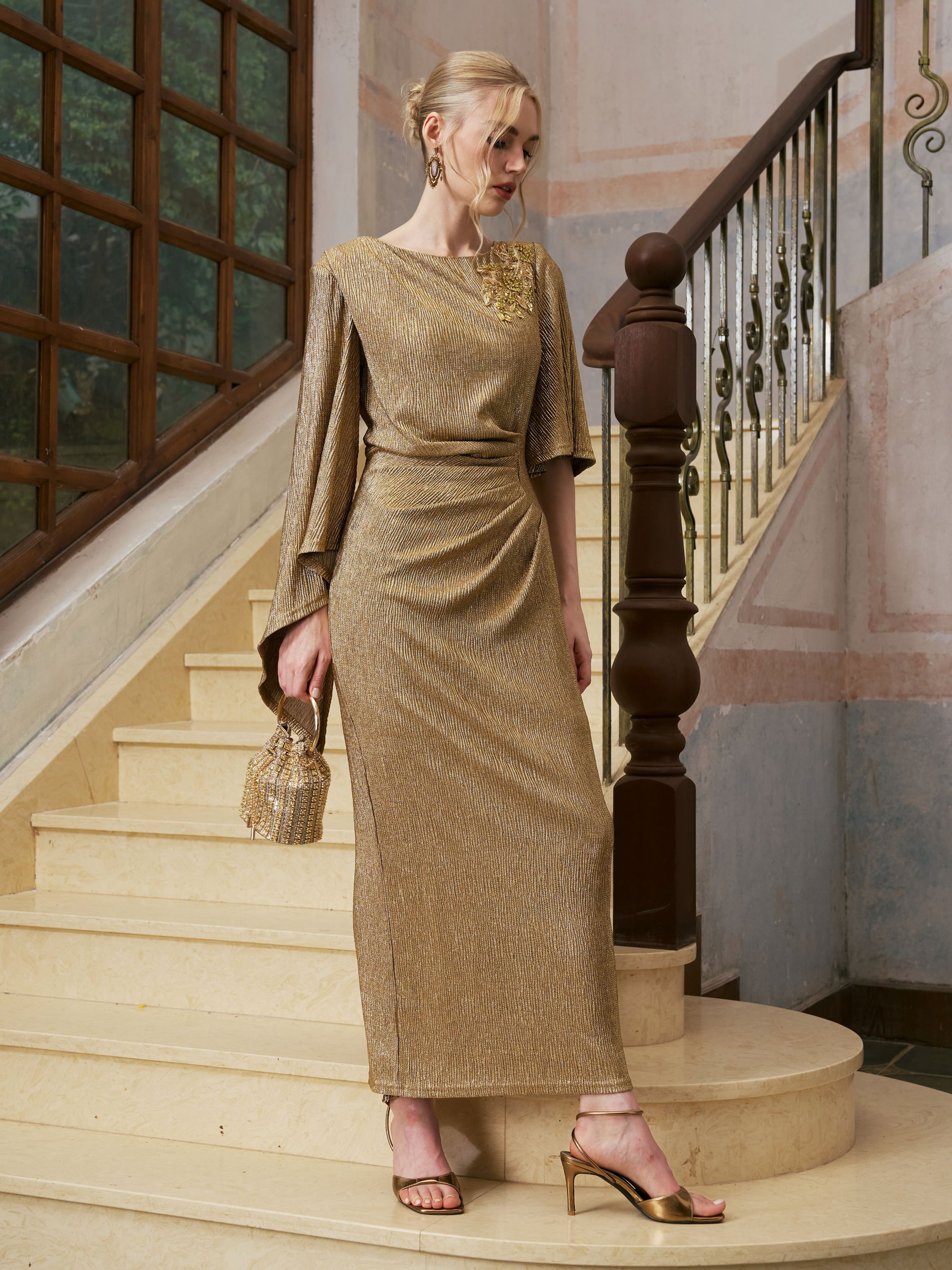 A woman wearing the Social Queen Floral Beaded Knit Dress, featuring draped details and gold accessories, stands on a staircase with a metal railing and large window.
