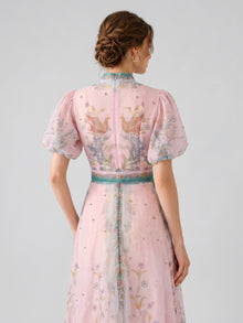 A woman with braided hair models the Social Queen Peacock Botanical Print Dress, a light pink A-line dress featuring puff sleeves, botanical and peacock motifs—including bird designs on the back—and a coordinating belt, set against a plain background.
